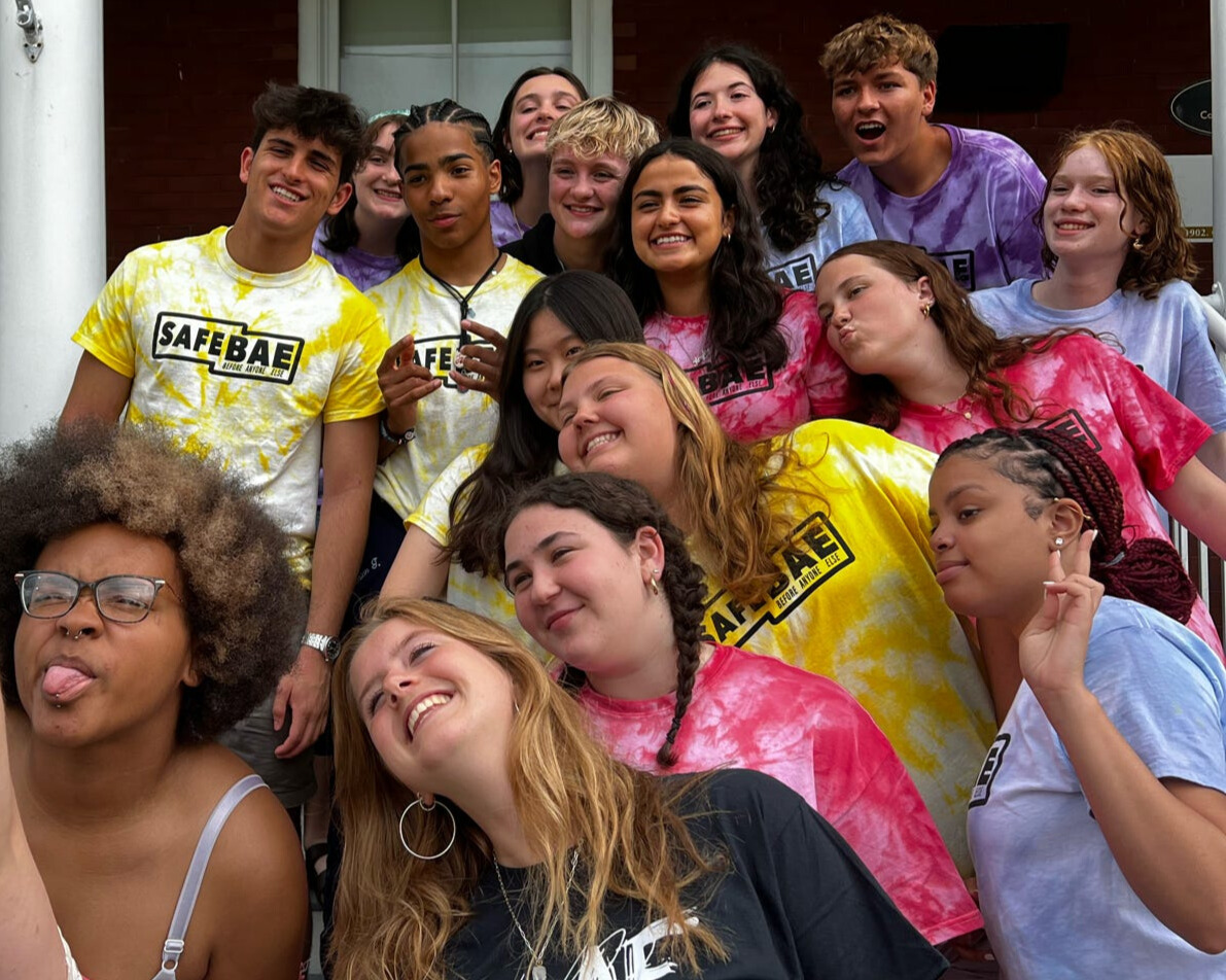 A group of SafeBAE student leaders in tie-dye SafeBAE shirts smiling and posing together on a set of steps.